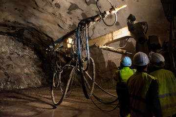 Back view of male engineers in uniform and helmets using drill equipment for boring passage during mining work underground