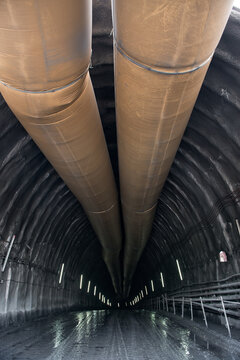 Wide passage in mining tunnel with glowing lamps on stone walls and huge metal pipe