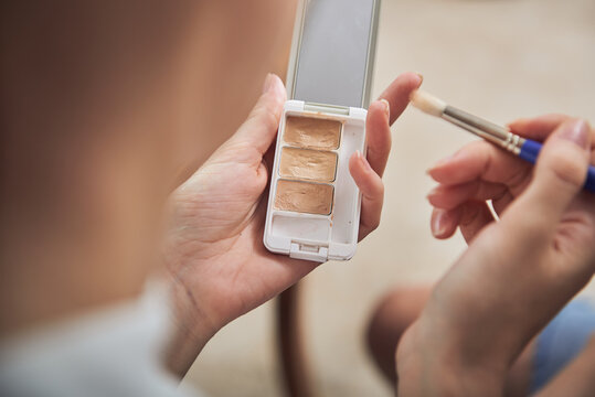 From behind crop anonymous female using soft makeup brush and palette of concealers while applying trendy makeup on face