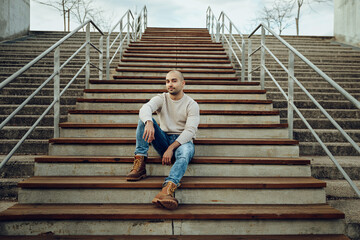 From below full length of positive young unshaven male in casual sweater and jeans with trendy boots sitting on stairway on street and looking at camera