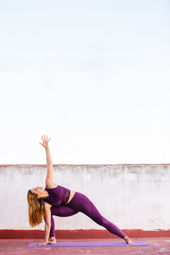 Side view of flexible female wearing bra and leggings practicing yoga in extended side angle pose on mat on terrace