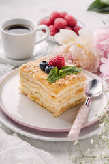 Slice cake of puff pastry with mint, raspberry and blueberry on plate, cup of coffee, peony. Delicate and airy composition, vertical format.