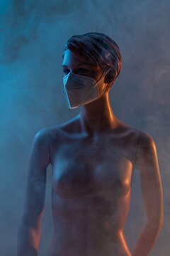 Plastic female mannequin in medical mask illuminated by colorful lights in studio with smoke on blue background