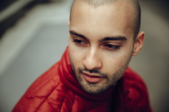 Young unshaven bald male in red warm jacket looking away with pensive smile against blurred outdoors background