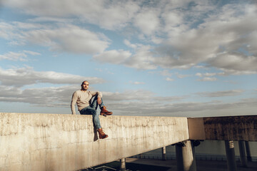 Side view of male in casual outfit sitting alone on border concrete structure against cloudy sky