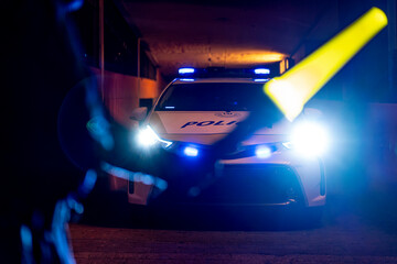 Back view of crop police officer standing with illuminated traffic wand in front of patrol car with flashing light at night