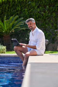 Side view of busy male entrepreneur sitting at poolside with legs in water and browsing netbook while working remotely on sunny day