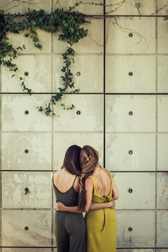 Back view of slender female friends with pigtail tenderly hugging on street on background of stone wall