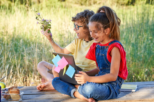 A ten year old boy and girl sitting at a wooden table in the field where there is a computer, flowers, fruit, snack. Dressed in denim and colorful T-shirts.