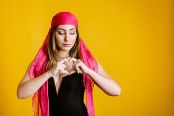 Peaceful female in headdress standing on yellow background in studio and demonstrating heart shape sign
