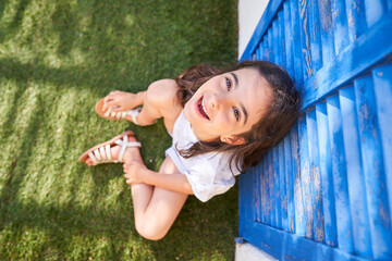 From above of happy carefree preteen girl looking up and smiling while sitting on green lawn and leaning against shabby blue shuttered window during summer day in countryside