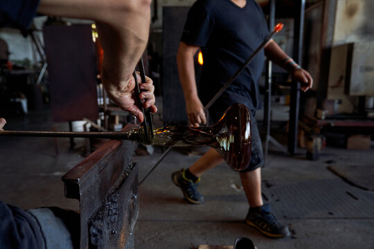 Crop men in workshop blowing glass base for hookah with blowpipe and tweezers