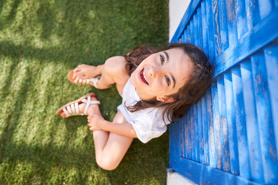 From above of happy carefree preteen girl looking up and smiling while sitting on green lawn and leaning against shabby blue shuttered window during summer day in countryside