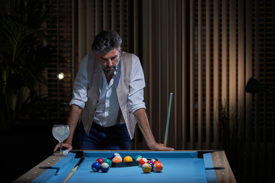 Thoughtful male in casual style standing with cue and thinking about game while leaning on pool table