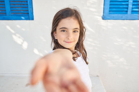 Adorable preteen girl outstretching hand forward camera and smiling while standing against white wall of country house in summer day