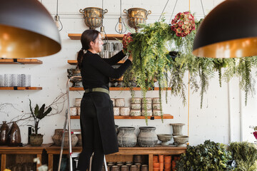Side view of female florist standing on ladder and arranging composition of green decorative hanging plants while working in cozy floristry salon