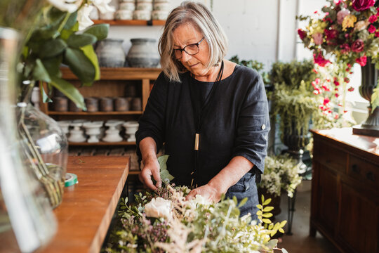 Professional adult female florist choosing green decorative plants for composition while working in cozy floristry studio