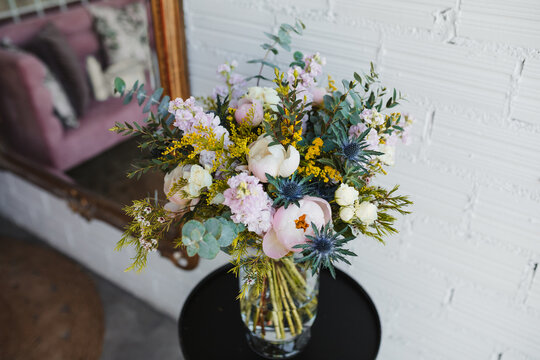 High angle of beautiful spring bouquet with assorted colorful blooming flowers including pink peonies and blue sea holly flowers with mimosa twigs arranged on glass vase on table near white wall