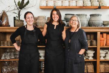 Positive professional female florists in black aprons smiling and looking at camera friendly while standing against shelves with various flower pots in stylish floristry shop