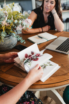 Crop anonymous female florist leafing through catalog of decorative plants while sitting at table with colleague and discussing new order for floristry workshop
