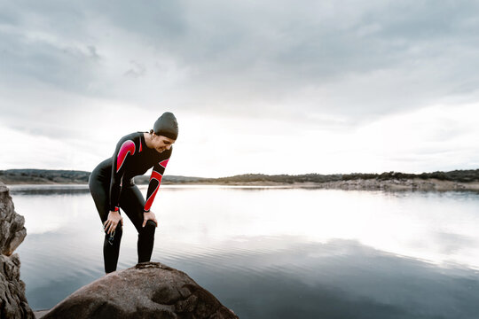 Side view slim tired female in black wetsuit standing resting on peaceful shore of lake in overcast