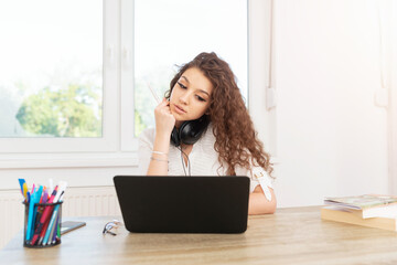 Pretty businesswoman in white shirt sitting in office by the window, using laptop and wearing headphones around neck.