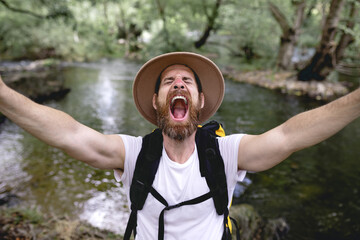 young man with beard hat on his head and yellow backpack hiking down a lake route with trees and shady areas taking a selfie