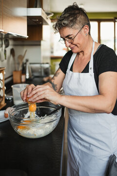 Smiling housewife in apron standing near table in modern kitchen and adding eggs in bowl with flour while preparing dough