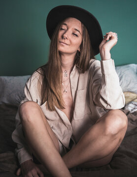 Stylish female in trendy hat and shirt sitting with crossed legs on soft bed and dreaming with closed eyes