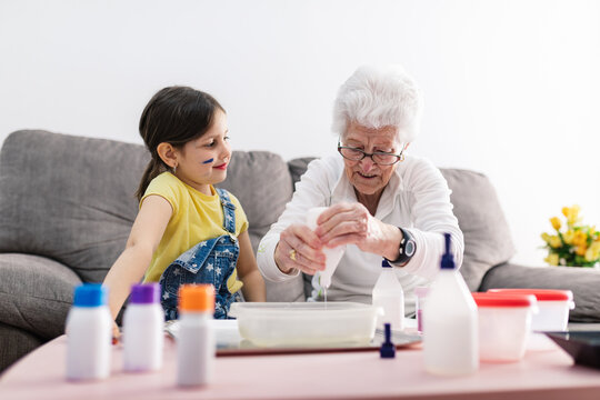 Smiling granny in eyeglasses helping positive granddaughter with dirty face in casual wear preparing blend using colorants in plastic container while sitting on comfortable couch in living room