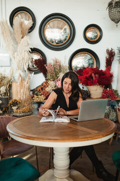 Positive adult female owner of stylish floristry salon sitting at wooden table with laptop and studying catalog of flowers while choosing plants for order
