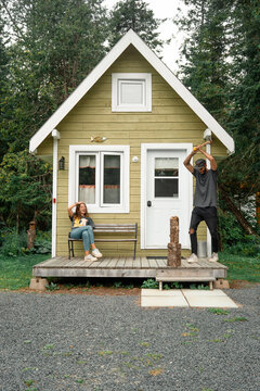 Woman chilling on bench on terrace of small rural house while man cutting wood nearby having weekend in New Brunswick