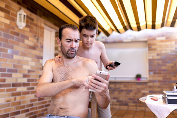 Confused shirtless bearded man surfing internet on cellphone while calm boy standing behind with trimmer in hairdressing salon near brick wall