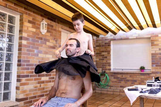 Serene shirtless teenager putting black wavy cape on adult father in jeans while sitting on stool in hairdressing salon near table with accessories