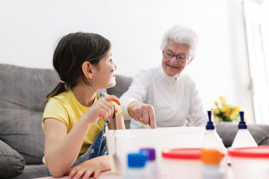 Smiling granny in eyeglasses helping positive granddaughter with dirty face in casual wear preparing blend using colorants in plastic container while sitting on comfortable couch in living room