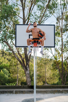 From below of male athlete in sportswear sitting with closed eyes meditating on basketball hoop with ball