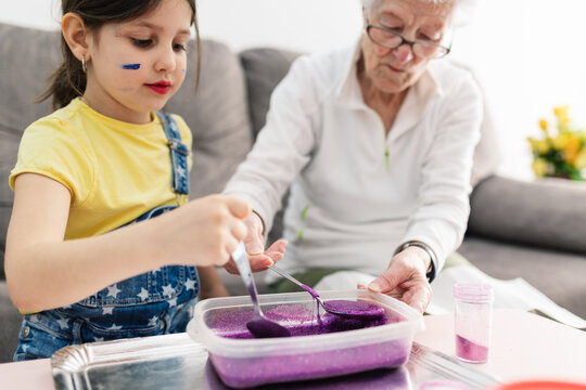 Smiling granny in eyeglasses helping positive granddaughter with dirty face in casual wear preparing blend using colorants in plastic container while sitting on comfortable couch in living room