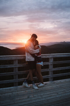 Full body hipster guy in cap embracing girlfriend while standing on wooden observation deck against ocean shore in beams of sundown