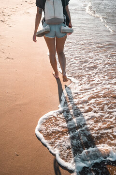 Crop pf back view of anonymous female tourist in summer clothes walking barefoot along sea during vacation