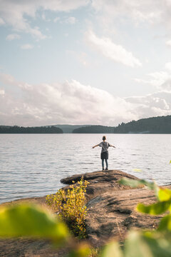 Back view of anonymous female hiker standing on stony shore near calm lake and enjoying freedom during cloudy summer day in Algonquin Provincial Park in Canada