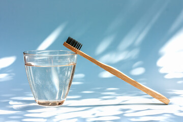 Bamboo toothbrush and glass of water on light blue pastel background with shadows of natural light. Biodegradable pesonal care and plastic free zero waste concept. Selective focus.