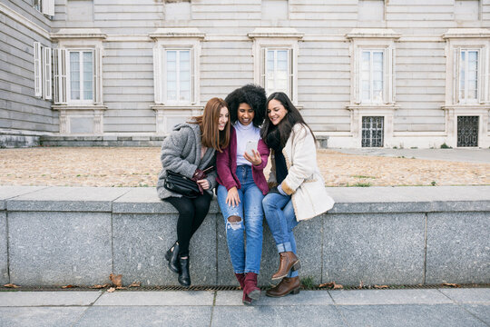 Female friends using smart phone while sitting on retaining wall against Madrid Royal Palace, Spain