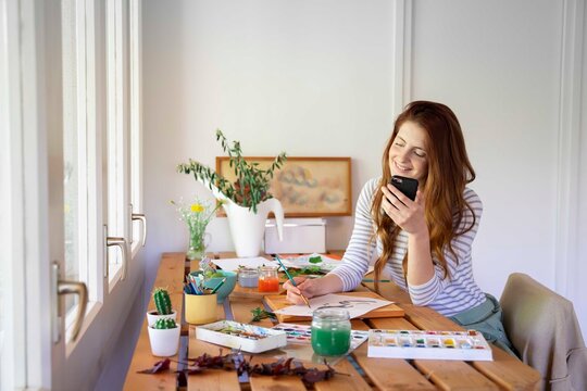 Happy young woman painting while holding smart phone on table at home