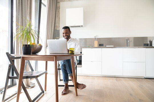 Portrait Of Smiling Man Sitting At Table In Modern Apartment Using Laptop And Earphones