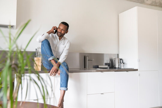 Portrait of serious man on the phone sitting on kitchen counter at home looking at distance