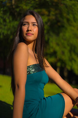 Naklejka premium A pretty young brunette Latina with long straight hair, wearing a tight green dress. Portrait of a serious young woman in a park in summer