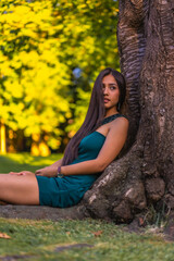 A young pretty Latina brunette with long straight hair leaning against a tree in a green dress. Sitting by a tree in the park