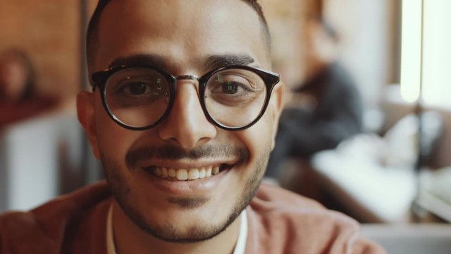 Close up portrait shot of young handsome middle eastern man in glasses and casual outfit sitting in cafe, looking at camera and happily smiling