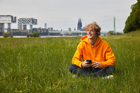 Happy young man sitting on a meadow listening music with headphones, Cologne, Germany - Powered by Adobe