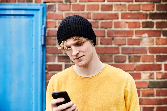Portrait of young man wearing cap looking at smartphone in front of brick wall
